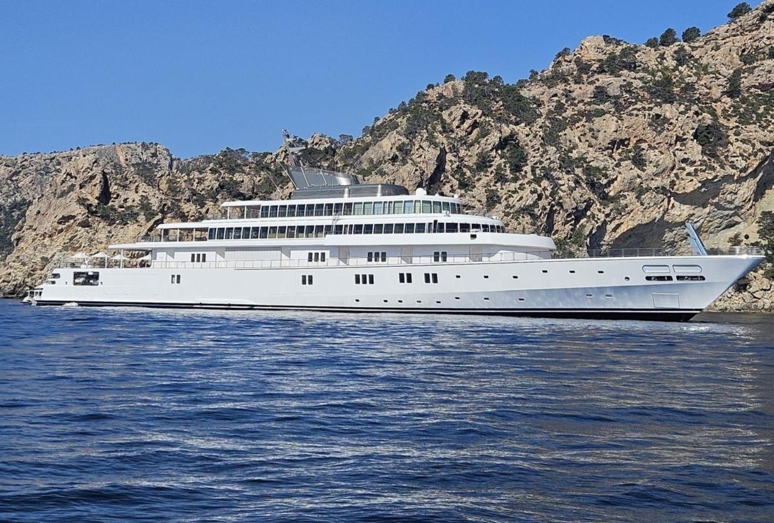Side profile of the 138-metre M/Y Rising Sun at anchor off a rocky coastline